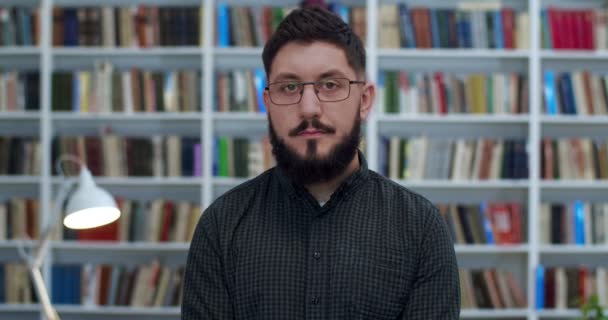 Retrato de un joven caucásico con barba en gafas mirando a la cámara con una leve sonrisa en la biblioteca. Primer plano del profesor masculino o trabajador de bibliotheca. Libros estantes en el fondo. Profesora principal . — Vídeo de stock