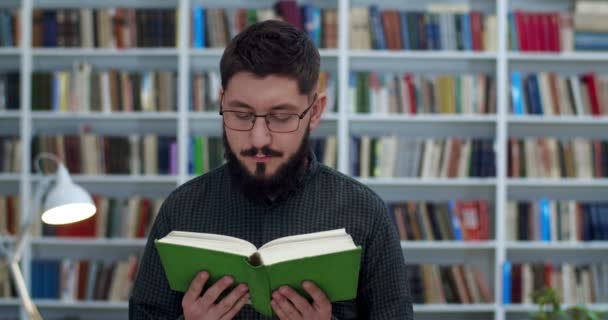 Närbild av unga kaukasiska stilig man i glasögon och med skägg läsa bok i biblioteket. Snygg manlig läsare med lärobok i händerna studerar i bibliotheca. Porträtt av en kille som ler mot kameran. — Stockvideo