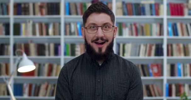 Portret van een knappe man in een bril die in de bibliotheek staat en glimlachend naar de camera kijkt. Close-up van vrolijke mannelijke resercher in bibliotheca glimlachend met verbaasd gezicht. Boeken planken op de achtergrond. — Stockvideo