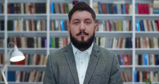 Young Caucasian businessman standing in cabinet with books shelves and looking straight to camera. Portrait of man with beard in public library or book store. Male professor. — Stock Video