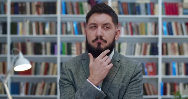 Jeune homme d'affaires caucasien toucher sa barbe et penser à quelque chose très soigneusement sur les étagères de livres derrière. Portrait d'un homme sérieux envisageant une théorie philosophique à la bibliothèque. Concept d'étude . — Video