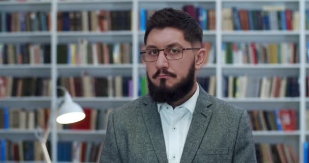 Porträtt av vit ung affärsman i glasögon som står i biblioteket, tittar på sidan och vänder ansikte mot kamera. En manlig smart lärare. Professor i bibliotheca. Man kund i bokhandeln. — Stockvideo