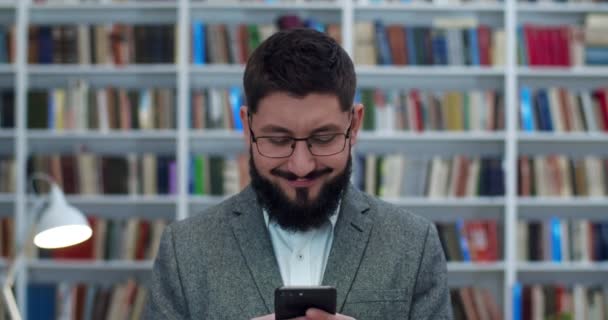 Souriant jeune homme caucasien dans des lunettes debout et tapotant sur smartphone dans la bibliothèque. Professeur ou chercheur masculin avec téléphone dans les mains textos message dans bibliotheca et souriant. Concept de discussion . — Video