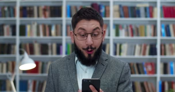 Kaukasischer junger gutaussehender Mann mit Brille und Bart blickt auf den Smartphone-Bildschirm und freut sich über gute Nachrichten. Männliche Bibliothekskunden lesen schöne Nachrichten am Telefon. Ja-Handbewegung. — Stockvideo