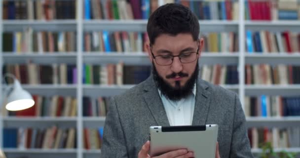 Kaukasisk ung stilig affärsman står i biblioteket och håller surfplatta enhet, knacka och bläddra. Man tittar på något på surfplatta dator. Manlig forskare i bibliotheca. Studieupplägg. — Stockvideo