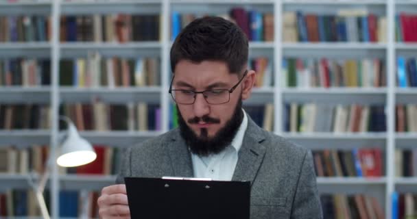 図書館に立っている眼鏡の白人の若い男性教授は、フォルダ内の文書と何かを注記し、書きます。ひげを生やした男、書誌の研究者そしていくつかの論文をチェックする. — ストック動画