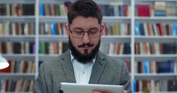 Gros plan de jeune homme d'affaires caucasien dans des lunettes debout dans la bibliothèque et tenant tablette dispositif, tapotant et défilant à l'écran. Un homme regardant quelque chose sur tablette. Chercheur masculin étudiant . — Video