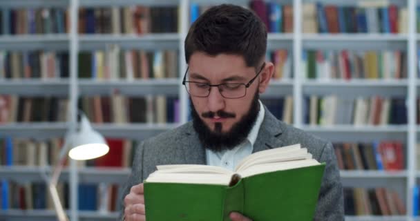 Joven hombre de negocios caucásico leyendo libro de texto en la sala de la biblioteca y estudiando. Retrato de hombre guapo en gafas cerrando libro y sonriendo a cámara en espacio bibliotheca . — Vídeo de stock