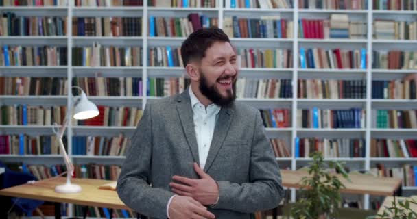 Портрет молодого щасливого кавказького чоловіка в куртці і з бородою сміються голосно перед камерою в бібліотечній кімнаті. Веселий чоловік з книжкового магазину посміхається і розважається. Бізнесмен у бібліотеці — стокове відео