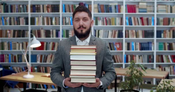 Portrait of young handsome Caucasian man with stack of textbooks standing in libary and smiling to camera. Male student in bibliotheca. Customer of book store. Study concept. — Stock Video