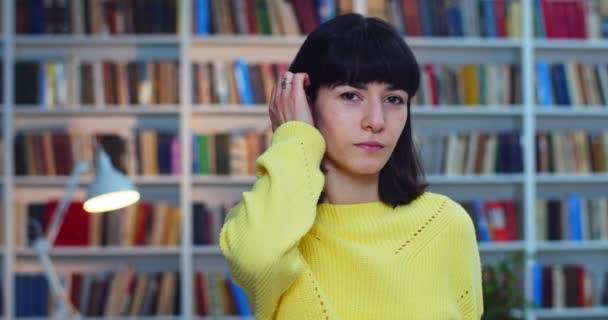 Nahaufnahme Porträt eines nachdenklichen brünetten Mädchens mit gelbem Pullover, das ihr Haar berührt und neben dem Bücherregal in der Bibliothek steht — Stockvideo