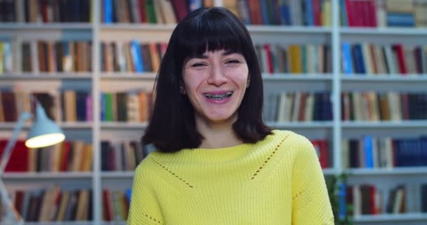 Zbliżenie portret uśmiechniętej brunetki z aparatem na zębach i żółtym swetrem stojącej blisko regału w bibliotece — Wideo stockowe