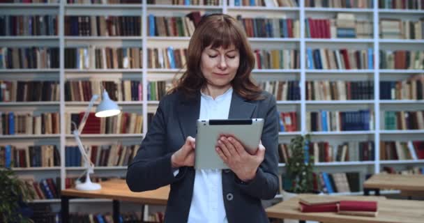 Zbliżenie portret kobiety architektki w okularach casual za pomocą tabletu w bibliotece i przygotowuje prezentację bissnes. Regał na książki w tle. — Wideo stockowe