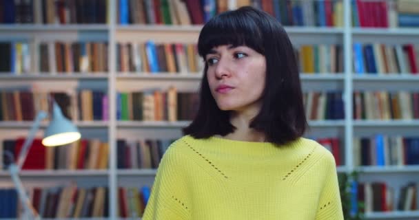 Крупним планом портрет вдумливої молодої жінки в жовтому светрі, що стоїть біля книжкової полиці в бібліотеці — стокове відео