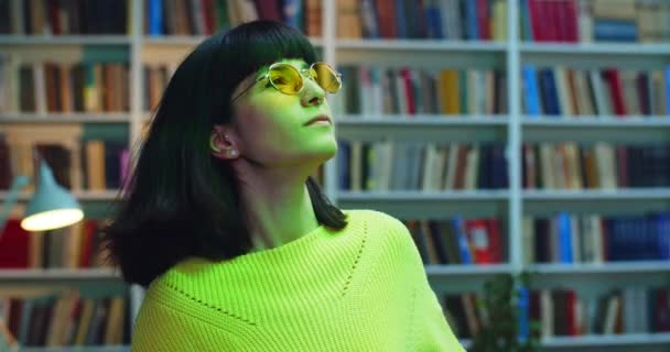 Retrato de cerca de una chica hipster bailando en gafas de sol amarillas y suéter contra la estantería de la biblioteca con luces de discoteca . — Vídeos de Stock