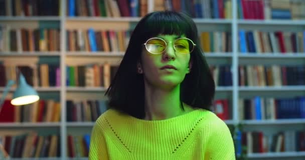 Großaufnahme Porträt einer tanzenden Brünette mit gelber Sonnenbrille und Pullover gegen Bücherregal in der Bibliothek mit bunten Neonlichtern. — Stockvideo