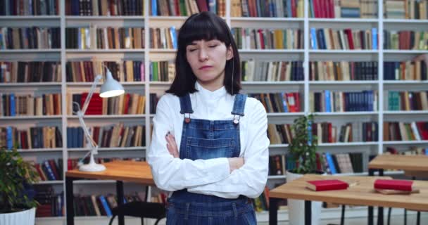 Close-up portret van een zelfverzekerde brunnete vrouw die in de bibliotheek staat en naar de camera kijkt. Boekenkast op de achtergrond. — Stockvideo