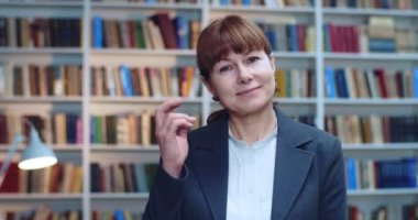 Kütüphanede kitaplığın yanında duran mutlu orta yaşlı kadın profesörün yakın plan portresi. Kameraya bakıyor ve saçına dokunurken gülümsüyor..