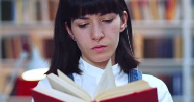 Esmer inek kızın yakın plan portresi son sınava hazırlanıyor ve kütüphanede kitap okuyor. Arkaplanda kitaplık.