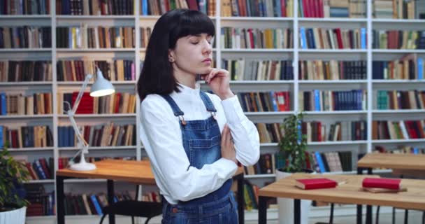 Ritratto ravvicinato di una giovane studentessa premurosa in piedi e che pensa in biblioteca. Libreria sullo sfondo . — Video Stock