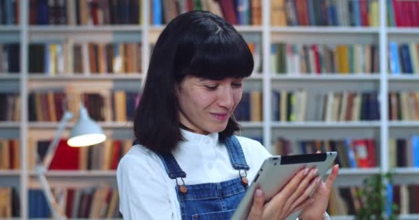 Close-up portret van een vrolijke studente in de bibliotheek met tablet en glimlach. Boekenkast op de achtergrond. — Stockvideo