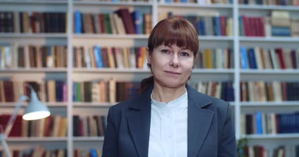 Close-up portret van volwassen profeshional wetenschapper met een bril in de bibliotheek naast boekenrek en kijkend naar de camera. — Stockvideo