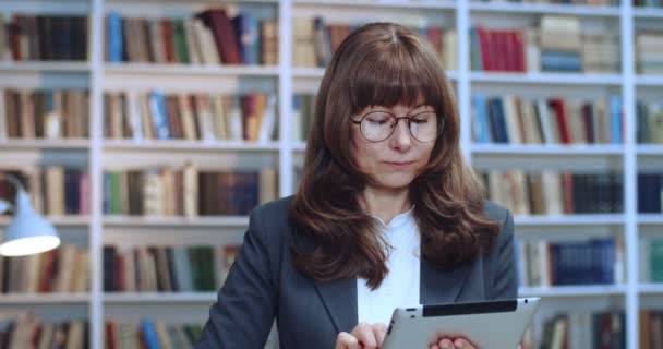 Zbliżenie portret kobiety architektki w okularach casual za pomocą tabletu w bibliotece i przygotowuje prezentację bissnes. Regał na książki w tle. — Wideo stockowe