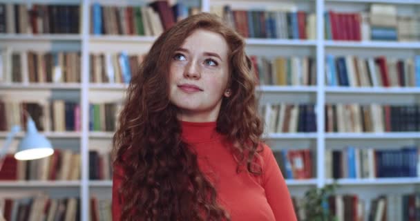 Retrato de una atractiva estudiante pelirroja con el pelo rizado largo y natural de pie en la biblioteca y sonriendo. Librería en segundo plano . — Vídeo de stock