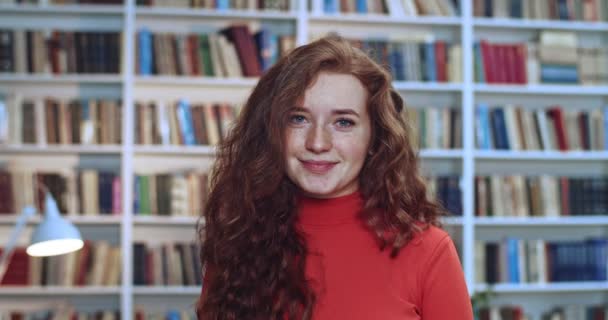 Портрет привлекательной рыжеволосой студентки с длинными вьющимися натуральными волосами, стоящей в библиотеке и играющей с волосами. Книжный шкаф в фоновом режиме . — стоковое видео