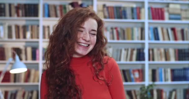 Портрет милой рыжей девушки с длинными вьющимися натуральными волосами, развлекающейся в библиотеке. Книжный шкаф в фоновом режиме . — стоковое видео