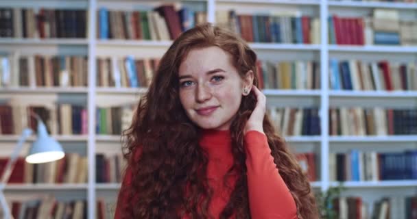 迷人的红头发、一头长长的卷曲自然的年轻女子站在图书馆里玩她的头发的画像。背景资料书柜. — 图库视频影像