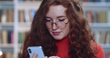 Kütüphanede siyah telefonunu kullanarak internette gezinen uzun kıvırcık saçlı, doğal çilli ve gözlüklü sevimli kızıl saçlı öğrencinin portresi. Arkaplanda kitaplık.