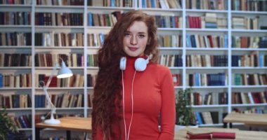 Uzun kıvırcık saç çilleri ve kütüphanede gülümseyen beyaz kulaklıkları olan sevimli kızıl saçlı öğrencinin portresi. Arkaplanda kitaplık.