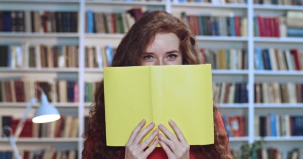 図書館や笑顔で楽しんでいる長い巻き毛の自然な髪と黄色の本を持つ遊び心のある赤い頭のかわいい女の子の肖像画。バックグラウンドの本棚. — ストック動画