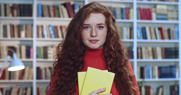 Portret van schattig rood hoofd lief meisje met lang krullend natuurlijk haar en gele boeken studeren in de bibliotheek. Boekenkast op de achtergrond. — Stockvideo