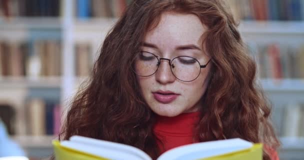 在图书馆里，一个有着一头长长的卷曲的自然发斑，戴着眼镜阅读黄皮书的漂亮红头发姑娘的画像。背景资料书柜. — 图库视频影像