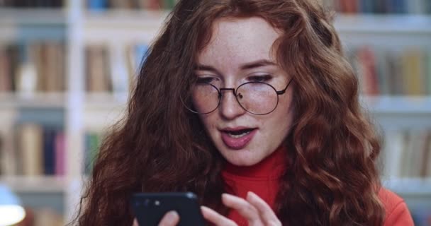 Ritratto di carina studentessa testa rossa con lunghe lentiggini capelli ricci naturali e occhiali sms con l'amico utilizzando il suo telefono nero in biblioteca. Libreria sullo sfondo . — Video Stock