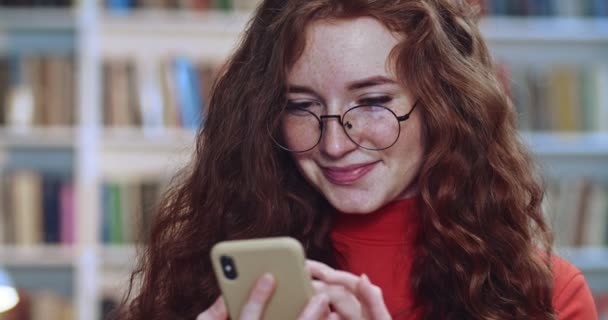 Porträtt av kaxig röd huvud flicka med långa lockigt naturliga hår fräknar och glasögon läser från sin gula telefon i biblioteket. Bokhylla i bakgrunden. — Stockvideo
