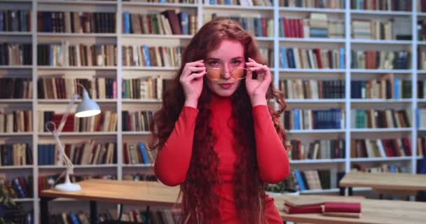 Porträt des süßen roten Kopfes freche Mädchen mit langen lockigen natürlichen Haaren Sommersprossen tragen gelbe Hipster-Sonnenbrille und tanzen in der Bibliothek mit Neonlicht. Bücherregal im Hintergrund. — Stockvideo