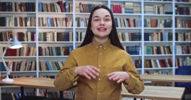 Retrato de feliz estudiante activa emocionalmente hablando y haciendo un montón de gestos por sus manos en la biblioteca . — Vídeo de stock