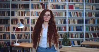 Gözlüklü mutlu kızıl saçlı kızın portresi kameraya doğru yürüyor ve kütüphanede kameraya gülümsüyor. Arkaplanda kitaplık.