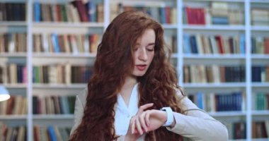 Kütüphanede akıllı saatini kullanan uzun kıvırcık saçlı kızıl saçlı genç bir kadının portresi. Arkaplanda kitaplık.