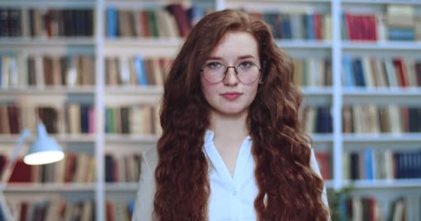 Close-up portret van aantrekkelijke roodharige jonge vrouw in een bril die glimlacht naar de camera. Boekenkast op de achtergrond. — Stockvideo