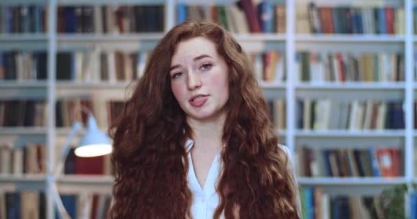 Porträt einer schönen jungen Studentin mit rotem Kopf, die in der Bibliothek emotional spricht und erklärt. Bücherregal im Hintergrund. — Stockvideo