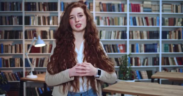 아름다운 빨간 머리 소녀가 감정적으로 말하고 도서관에서 손으로 많은 정보를 수집하는 모습. 배경의 북 케이스. — 비디오