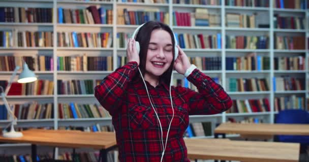 Ritratto di dolce bella studentessa bruna con fantastici capelli lunghi naturali e cuffie bianche che ballano e si divertono in biblioteca. Libreria sullo sfondo . — Video Stock