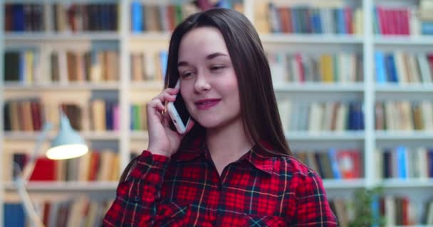 Portrait of friendly happy girl in checked shirt speaking at mobile phone in library. — Stock Video