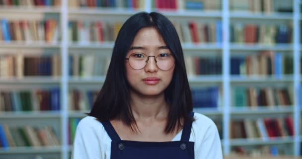 Retrato de hermosa asiática adolescente estudiante con pelo negro y gafas mirando a la cámara y sonriendo en la biblioteca al lado de estante de libros . — Vídeo de stock