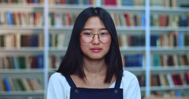 Siyah saçlı ve gözlüklü mutlu Asyalı genç kızın portresi. Kameraya bakıyor ve kütüphanede kitaplığın yanında uzanıyor.. — Stok video