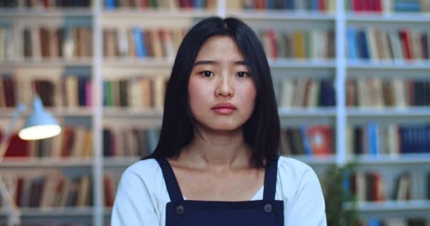 Retrato de estudiante adolescente asiática reflexiva con el pelo negro mirando a la cámara y pensando en el examen en la biblioteca al lado del estante del libro . — Vídeo de stock